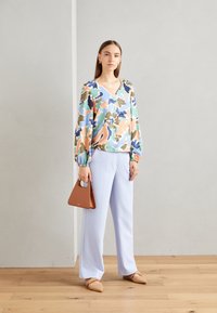 Blusa multicolore con un motivo floreale blu, verde e pesca, abbinata a pantaloni larghi azzurri e una piccola borsa marrone.
