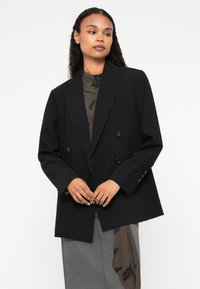 Blazer nero doppiopetto con design su misura, revers a punte e dettagli con bottoni, abbinato a un top marrone a fantasia e una gonna grigia.