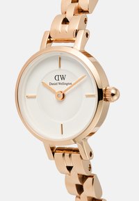 Apļa zelta toņa pulkstenis ar baltu ciparnīcu, minimālistiskām atzīmēm un savienotu rozā zelta aproci. Izceļas ar zīmola nosaukumu "Daniel Wellington."