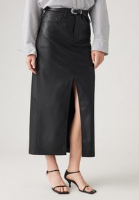 Jupe maxi en cuir noir avec une fente à l'avant, des passants de ceinture et des détails cousus ; associée à des sandales à talons hauts noires à lanières.