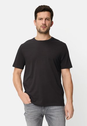 Mann mit kurzen dunklen Haaren und Bart, trägt ein schlichtes schwarzes kurzärmliges T-Shirt und graue Jeans, steht mit einer Hand in der Tasche vor weißem Hintergrund.