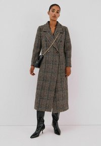 Dubbelzijdige, knielange jas van bruine geruite stof met een oversized kraag. Gecombineerd met zwarte, puntige laarzen en een kleine handtas.
