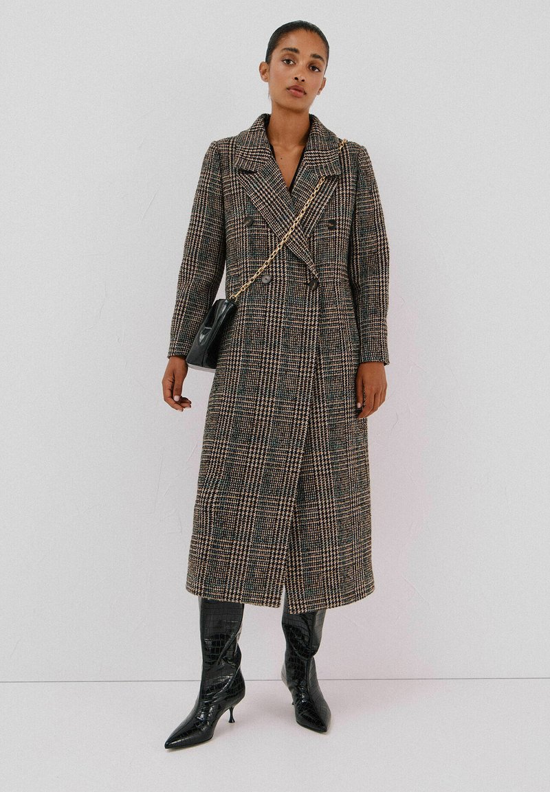 Dubbelzijdige, knielange jas van bruine geruite stof met een oversized kraag. Gecombineerd met zwarte, puntige laarzen en een kleine handtas.