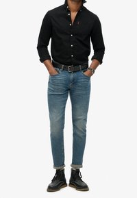 Camicia nera con bottoni, jeans blu slim-fit e stivali neri in pelle con lacci. Design semplice con maniche arrotolate e cintura in pelle.