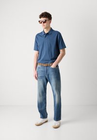 Ναυτικό μπλε πόλο μπλουζάκι με κοντά μανίκια, ανοιχτό μπλε ξεθωριασμένο τζιν, καφέ ζώνη και λευκά αθλητικά παπούτσια με μπεζ λεπτομέρειες. Τετράγωνα γυαλιά ηλίου.