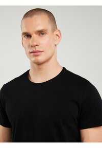 Jeune homme avec une coupe courte et les yeux bleus, portant un t-shirt noir à col rond, sur un fond gris clair.