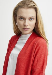 Röd cardigan med öppen front, texturerad stickning, långa ärmar och avslappnad passform, matchad med en vit hög-halsad topp.