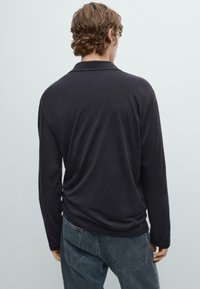 Persona con capelli corti e mossi che indossa una maglietta nera a maniche lunghe e jeans blu sbiaditi, in piedi di spalle contro uno sfondo neutro.