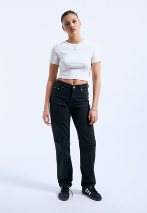 Mladá žena stojí, oblečená v bílém krátkém tričku, černých džínech s vysokým pasem, černých teniskách a stříbrném šperku na jednobarevném pozadí.