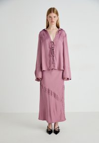Abito di seta rosa con maniche lunghe, dettagli in pizzo sulle spalle e sulla gonna, scollatura con cordoncino e un accento in pizzo diagonale. Tacchi neri.