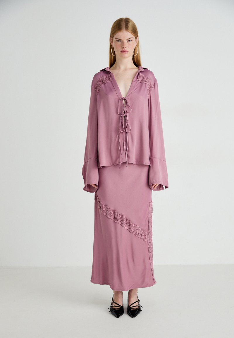 Abito di seta rosa con maniche lunghe, dettagli in pizzo sulle spalle e sulla gonna, scollatura con cordoncino e un accento in pizzo diagonale. Tacchi neri.