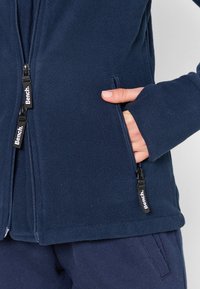 Giacca in pile blu navy con tasche con cerniera, che mette in mostra una morbida texture e un colletto arrotondato. Presenta tirazip neri con il logo 'Bench'.