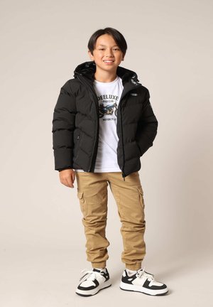 Doudoune noire avec capuche, fermeture éclair à l'avant et poignets élastiques. Portée par-dessus un t-shirt graphique blanc, associée à un pantalon cargo beige et des baskets.