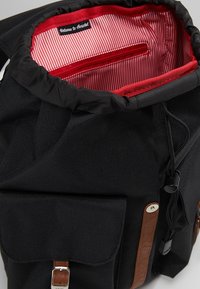 Zwarte stoffen rugzak met een rood-wit gestreepte binnenkant, trekkoordsluiting en bruine leren accenten; voorzien van een voorvak en een ritssluiting.