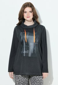 Blusa negra de manga larga con cuello vuelto, que presenta un estampado gráfico y cordones naranjas. Combinada con pantalones de patrón gris y negro.