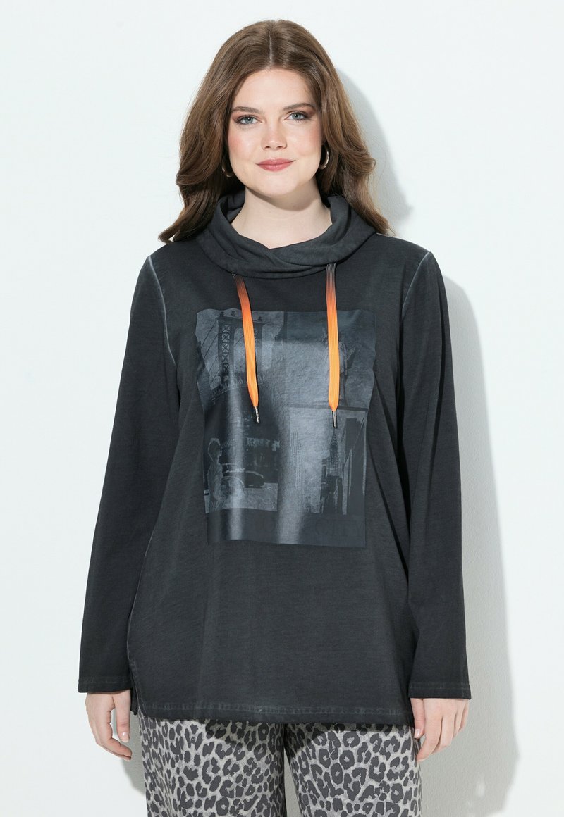 Blusa negra de manga larga con cuello vuelto, que presenta un estampado gráfico y cordones naranjas. Combinada con pantalones de patrón gris y negro.
