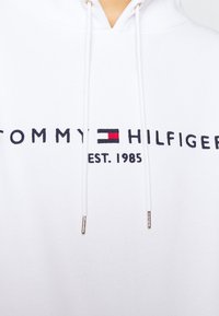 Sweat à capuche blanc avec un logo sur le devant indiquant "TOMMY HILFIGER EST. 1985." Accents noirs et rouges, avec cordons et extrémités en argent.