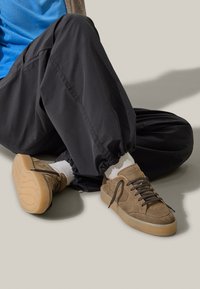 Sneaker in suede marrone con finitura texturizzata, suola piatta in gomma e lacci marroni scuri, abbinati a pantaloni neri a sigaretta e calze bianche.