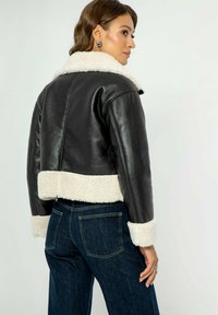 Giubbotto cropped in pelle nera con colletto e polsini in finta shearling color crema. Presenta una texture liscia e un design semplice. Indossato con denim scuro.