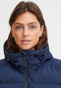 Tamnoplava puffer jakna s visokim ovratnikom, podesivim vezicama i zatvaračem sprijeda. Ima suptilan logotip na prsima.