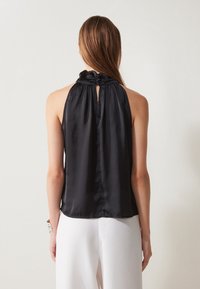 Černý saténový halter top s nabíraným výstřihem a otvorem na zádech. Hladká textura a volný střih. Stylizováno s bílými kalhotami.