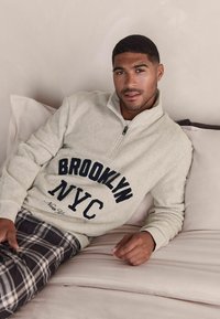 Lys grå fleece-pullover med "BROOKLYN NYC" tekst i mørk blå, kvart lynlås og ribbede manchetter, parret med sorte og hvide ternede bukser.