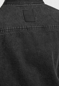 Vue arrière d'une veste en denim noire avec un empiècement cousu sur les épaules et un patch rectangulaire en cuir noir près du col.