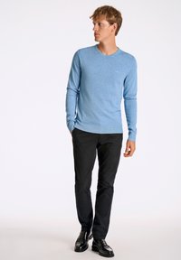 Vaaleansininen pitkähihainen neule, sukkamainen malli, pyöreä pääntie, valmistettu pehmeästä kankaasta. Yhdistetty mustiin housuihin ja kiiltäviin mustiin kenkiin.