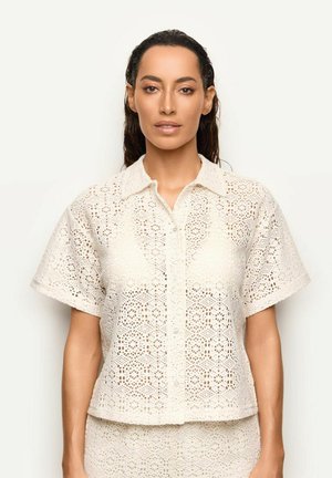 Donna con capelli scuri indossa un top crema all'uncinetto con maniche corte e bottoni e una gonna abbinata, in piedi davanti a uno sfondo chiaro e semplice.