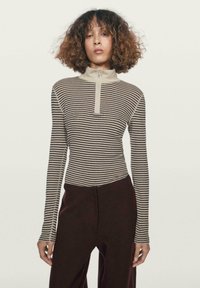 Beżowo-brązowa bluzka w paski z długim rękawem, z wysokim kołnierzem na pół suwaka, dopasowany fason i miękka faktura. Połączona z ciemnymi spodniami.