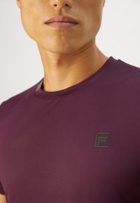 Homme portant un t-shirt de couleur prune avec un petit logo "F" noir et gris sur la poitrine gauche, sur fond uni.