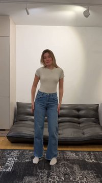 Jeune femme debout dans un salon, portant un t-shirt beige, un jean bleu, des chaussettes blanches, avec un canapé en cuir noir et un tapis à motifs derrière elle.