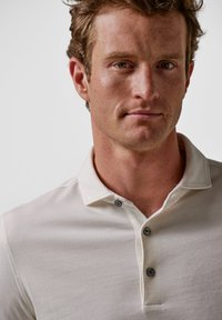 Ecrufarbenes Poloshirt mit Kragen und drei Knöpfen, aus weichem Stoff mit glatter Textur. Nahaufnahme des Oberkörpers.