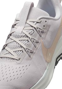 Lys grå sneaker med åndbart mesh-overdel, teksturerede accenter, subtil pink swoosh-logo og forstærket tåområde. Har en let sål.