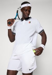 Jugador de tenis masculino con ropa deportiva blanca sostiene la raqueta sobre el hombro y coloca una pelota de tenis en el bolsillo de los pantalones cortos.