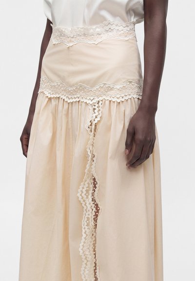 Mannequin à la peau foncée portant une jupe beige transparente avec des détails en dentelle et volants, associée à un haut blanc en soie, mains détendues le long du corps.