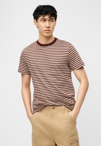 Νέος άνδρας με σκούρα σγουρά μαλλιά που φοράει κοντομάνικο ριγέ μπλουζάκι σε μπορντό και μπεζ και χακί παντελόνι cargo, με τα χέρια στις τσέπες.