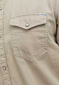 Camisa beige de botón fabricada en una mezcla de algodón, con un bolsillo en el pecho con tapa, botones a presión plateados y detalles de costura. Etiqueta de la marca visible.