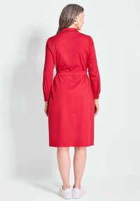 Rotes Hemdkleid mit einem taillierten Bund, langen Ärmeln und Knopfmanschetten. Glattes Material, knielanger Saum, getragen mit weißen Sneakers. Rückansicht.