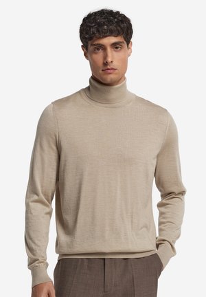 Homme portant un pull à col roulé beige et un pantalon marron, debout avec une main dans la poche devant un fond uni.