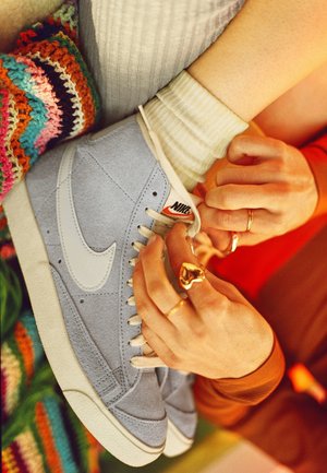 Mains avec des bagues en or nouant les lacets de baskets Nike bleu clair portées avec des chaussettes crème, à côté d'un tissu crocheté multicolore.