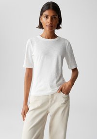 Donna con i capelli corti e scuri, indossa una maglietta bianca a maniche corte e pantaloni off-white, in piedi con una mano in tasca su uno sfondo semplice.