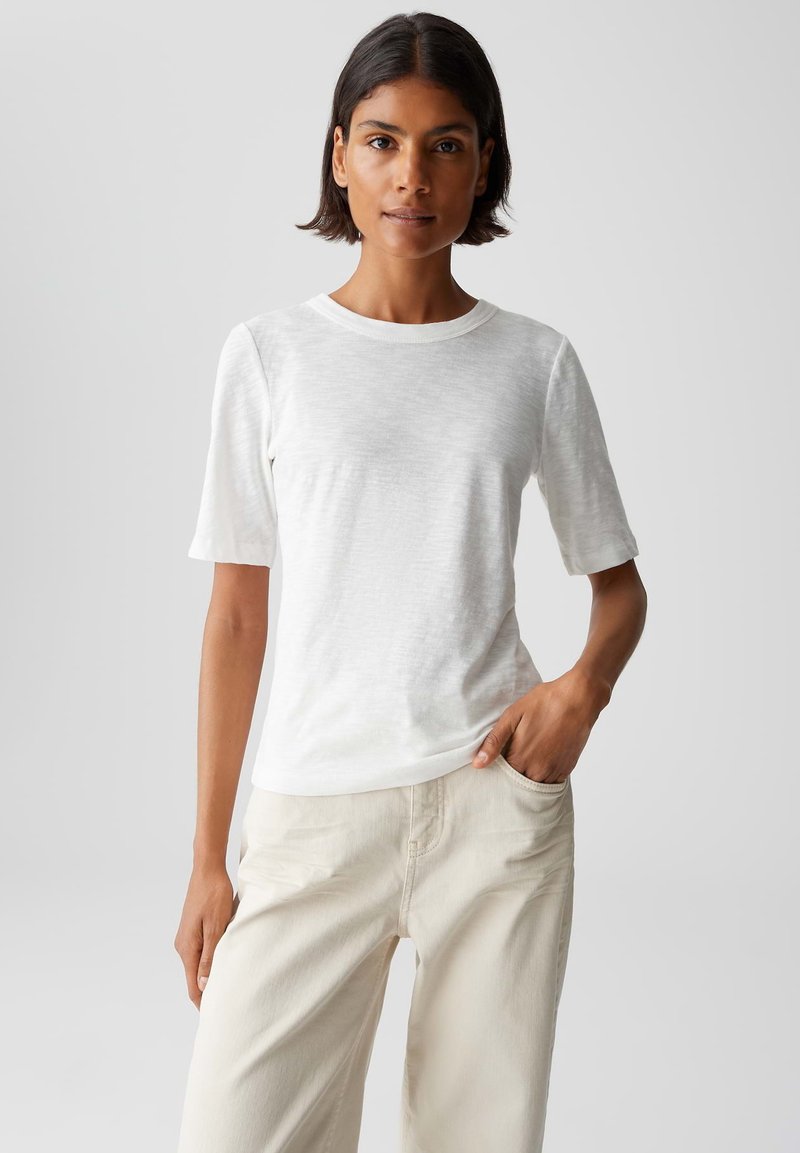 Donna con i capelli corti e scuri, indossa una maglietta bianca a maniche corte e pantaloni off-white, in piedi con una mano in tasca su uno sfondo semplice.