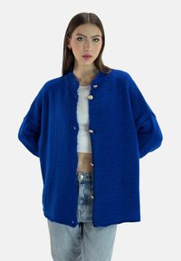 Cárdigan de punto azul brillante con escote redondo, que presenta grandes botones plateados y mangas oversize. Llevado sobre una blusa blanca y vaqueros.