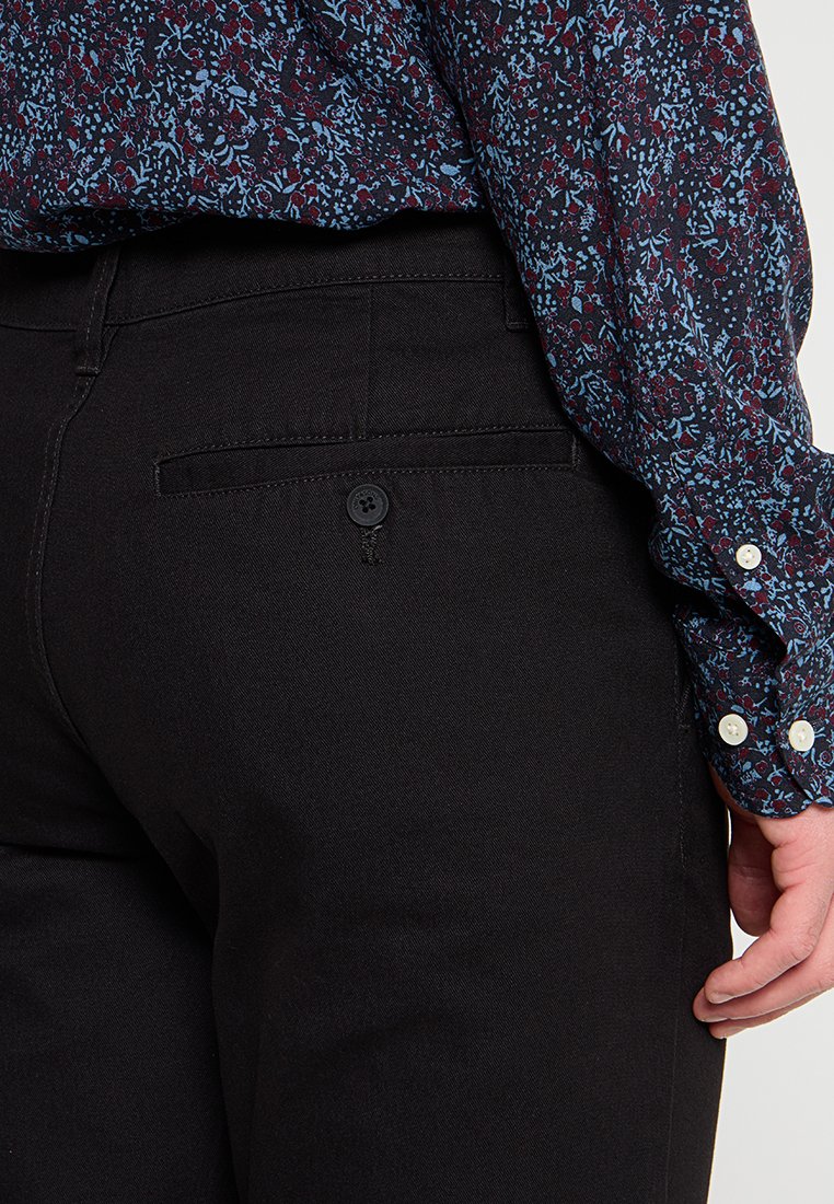 Person, der schwarze Hosen mit einer mit Knopf verschlossenen Gesäßtasche und ein langärmliges, floral gemustertes Shirt in Blau und Rot trägt.