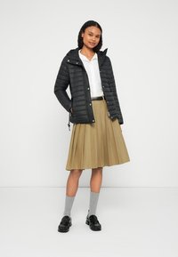 Kobieta ubrana w czarną kurtkę puchową, białą koszulę, beżową plisowaną spódnicę, szare skarpetki i czarne mokasyny, stojąca na tle jednolitego białego tła.