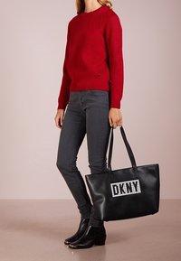 Černá taška přes rameno s mřížkovým vzorem a bílým logem "DKNY". Hladká kožená textura s dvojitými ramenními popruhy. Červený žebrovaný svetr.
