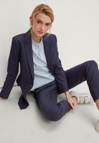 Blazer blu navy e pantaloni abbinati, indossati con una camicia a righe blu chiaro. Il modello è seduto su uno sfondo neutro, indossando sneakers bianche.