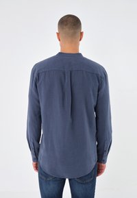 Uomo con capelli corti che indossa una camicia a maniche lunghe blu navy e jeans blu, rivolto di spalle contro uno sfondo bianco semplice.