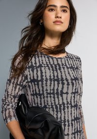 Langärmliges, leichtes Oberteil in Grau mit einem schwarzen abstrakten Muster. Das Modell hält eine schwarze Lederhandtasche, sie hat einen runden Ausschnitt und eine taillierte Passform.